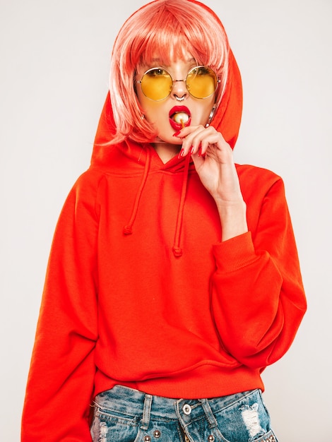 Retrato de joven hermosa hipster chica mala en moda verano rojo con capucha y arete en la nariz. Mujer rubia sonriente despreocupada sexy posando en el estudio con peluca. Modelo positivo lamiendo dulces de azúcar redondo