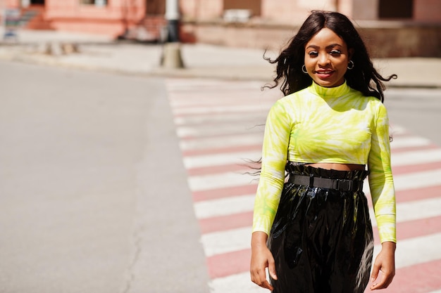 Retrato de una joven y hermosa modelo afroamericana caminando con ropa de cruce de peatones en top verde y pantalones negros brillantes de aspecto húmedo