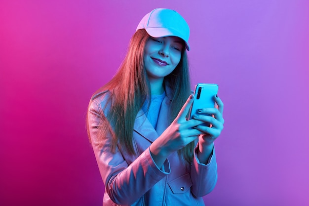 Retrato de joven hermosa modelo de moda con panadero de cuero y gorra de béisbol, sosteniendo el teléfono inteligente en las manos