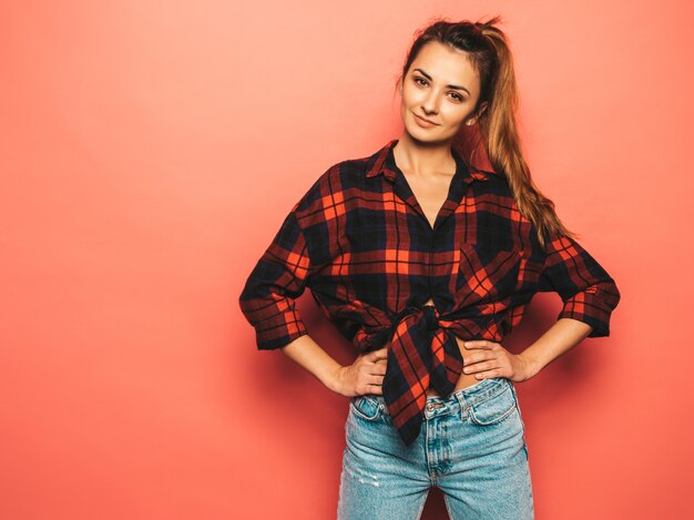 Retrato de joven hermosa muchacha sonriente hipster en ropa de jeans y camisa a cuadros de moda verano. Mujer despreocupada atractiva que presenta cerca de la pared rosada en estudio. Modelo positivo sin maquillaje.