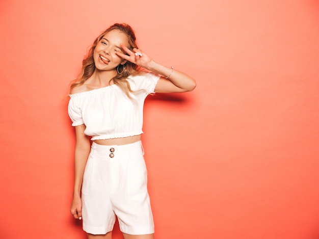 Retrato de joven hermosa muchacha sonriente hipster en ropa de moda de verano. Mujer despreocupada atractiva que presenta cerca de la pared rosada. Modelo positivo divirtiéndose Muestra el signo de pece y la lengua