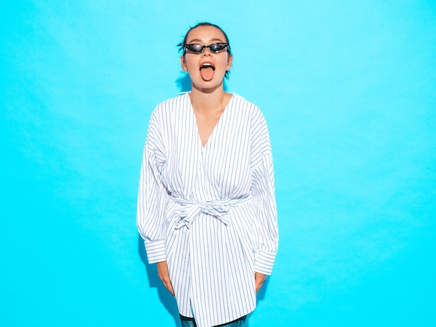 Retrato de joven hermosa muchacha sonriente hipster en ropa de moda de verano. Mujer despreocupada sexy posando junto a la pared azul. Modelo positivo divirtiéndose en gafas de sol. Muestra lengua