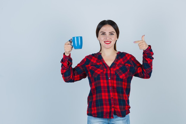 Retrato de joven hermosa mujer apuntando a la taza en camisa casual, jeans y mirando alegre vista frontal