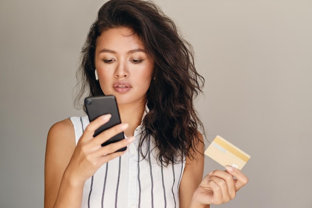 Retrato de una joven y hermosa mujer asiática con tarjeta de crédito usando un celular con confianza sobre un fondo gris