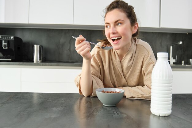 Retrato de una joven hermosa mujer en bata de baño comiendo cereales para el desayuno se apoya en la encimera de la cocina