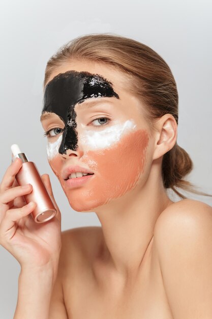 Retrato de una joven hermosa mujer con diferentes máscaras cosméticas en la cara sosteniendo un líquido de belleza en la mano mientras mira soñadoramente a la cámara sobre un fondo gris