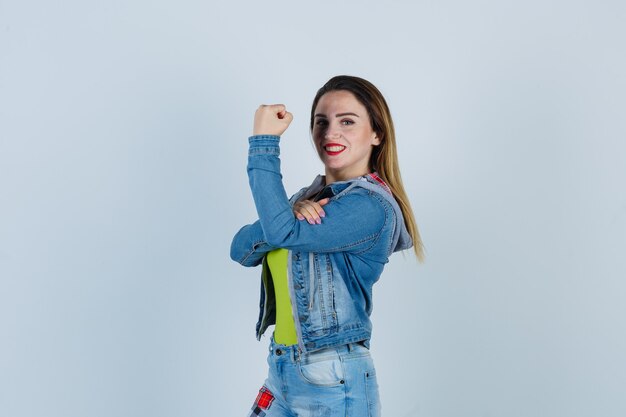 Retrato de joven hermosa mujer manteniendo la mano sobre los músculos del brazo en traje de mezclilla y mirando orgullosa vista frontal