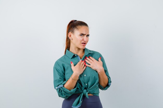 Retrato de joven hermosa mujer manteniendo las manos sobre el pecho en camisa verde y mirando indeciso vista frontal