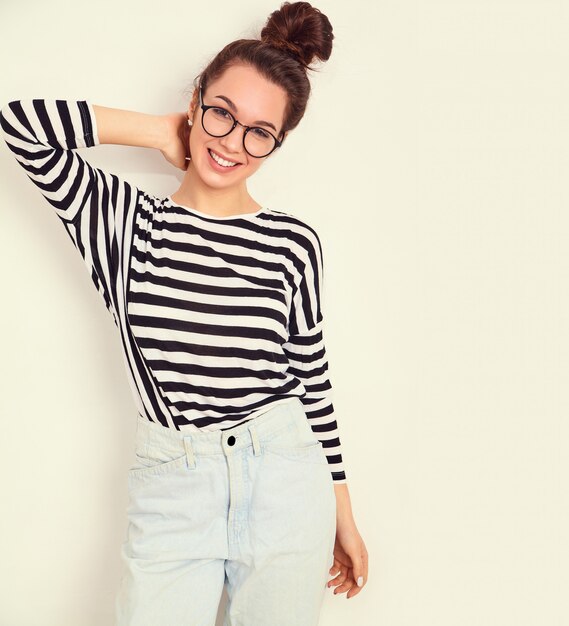 Retrato de joven hermosa mujer morena modelo chica con maquillaje desnudo en gafas en ropa hipster de verano posando junto a la pared.