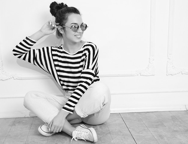 Retrato de joven hermosa mujer morena modelo chica con maquillaje desnudo en ropa hipster de verano posando junto a la pared en gafas de sol. sentado en el suelo