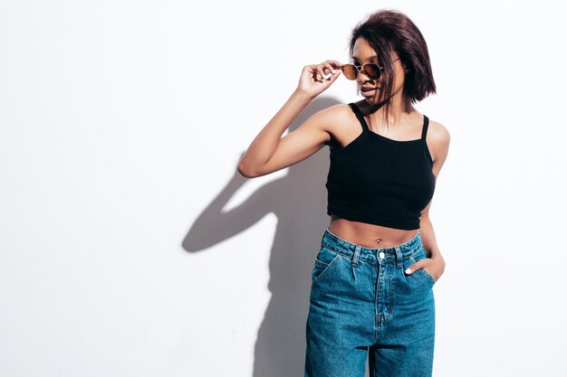 Retrato de joven hermosa mujer negra Modelo sonriente vestida con jeans de verano y ropa superior negra Sexy mujer despreocupada posando junto a la pared blanca en el estudio Bronceada y alegre