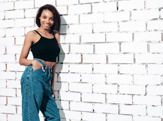 Retrato de joven hermosa mujer negra Modelo sonriente vestida con jeans de verano y ropa superior negra Sexy mujer despreocupada posando junto a la pared de ladrillo blanco en el estudio Bronceada y alegre