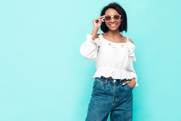 Retrato de joven hermosa mujer negra Modelo sonriente vestida con ropa de jeans de verano Sexy mujer despreocupada posando junto a la pared azul en el estudio Bronceada y alegre En gafas de sol