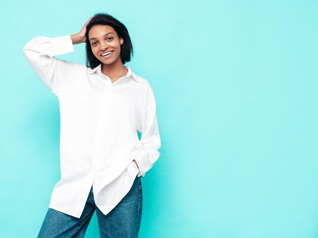 Retrato de joven hermosa mujer negra Modelo sonriente vestida con ropa de jeans de verano Sexy mujer despreocupada posando junto a la pared azul en el estudio Bronceada y alegre