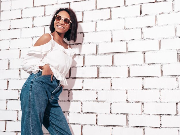 Retrato de joven hermosa mujer negra Modelo sonriente vestida con ropa de jeans de verano Sexy mujer despreocupada posando junto a la pared de ladrillo blanco en el estudio Bronceada y alegre En gafas de sol