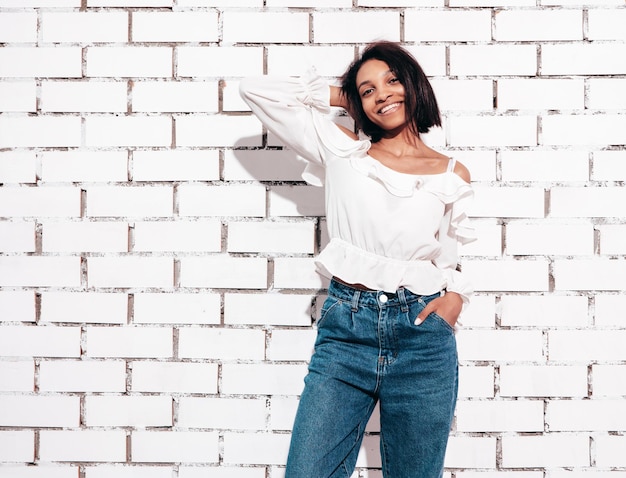 Retrato de joven hermosa mujer negra Modelo sonriente vestida con ropa de jeans de verano Sexy mujer despreocupada posando junto a la pared de ladrillo blanco en el estudio Bronceada y alegre