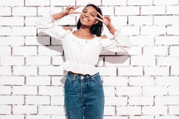 Foto gratuita retrato de joven hermosa mujer negra modelo sonriente vestida con ropa de jeans de verano sexy mujer despreocupada posando junto a la pared de ladrillo blanco en el estudio bronceada y alegre