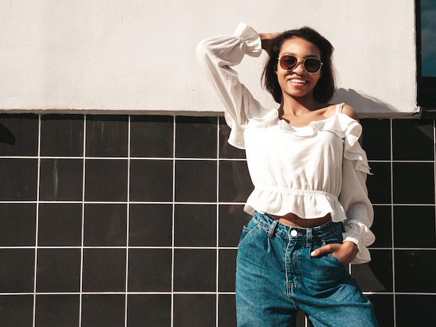 Retrato de joven hermosa mujer negra Sonriente modelo hipster en ropa de jeans blancos Sexy mujer despreocupada posando en el fondo de la calle Alegre y feliz al aire libre En gafas de sol Al atardecer