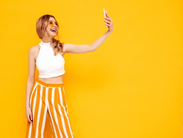 Retrato de joven hermosa mujer rubia sonriente en ropa de verano de moda mujer despreocupada posando cerca de la pared amarilla en el estudio Modelo positivo divirtiéndose en el interior Alegre y feliz Tomar foto selfie