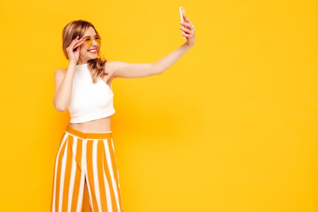 Foto gratuita retrato de joven hermosa mujer rubia sonriente en ropa de verano de moda mujer despreocupada posando cerca de la pared amarilla en el estudio modelo positivo divirtiéndose en el interior alegre y feliz tomar foto selfie