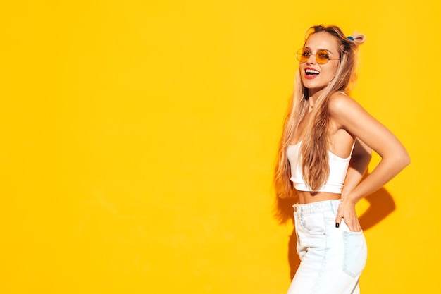 Retrato de joven hermosa mujer rubia sonriente en ropa de verano de moda Mujer despreocupada sexy posando junto a la pared amarilla en el estudio Modelo positivo divirtiéndose en el interior Alegre y feliz en gafas de sol