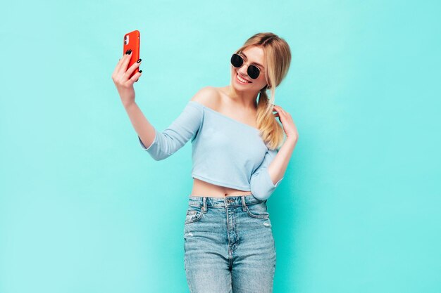 Retrato de joven hermosa mujer rubia sonriente en ropa de verano de moda Mujer despreocupada sexy posando junto a la pared azul en el estudio Modelo positivo divirtiéndose en el interior Alegre y feliz Tomando selfie
