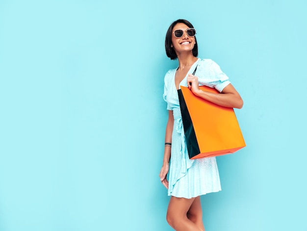 Retrato de joven hermosa mujer sonriente en vestido de verano de moda Mujer despreocupada posando cerca de la pared azul en el estudio Modelo positivo sosteniendo bolsa de compras Alegre y feliz Aislado