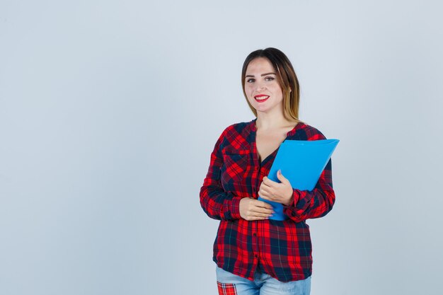 Retrato de joven hermosa mujer sosteniendo la carpeta, mirando al frente en camisa casual, jeans y mirando dichosa vista frontal