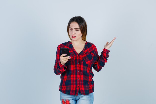 Retrato de joven hermosa mujer sosteniendo el teléfono, levantando la mano de manera interrogante en camisa casual y mirando molesto vista frontal