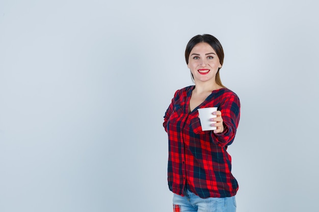Retrato de joven hermosa mujer sosteniendo un vaso de plástico mientras posa en camisa casual, jeans y mirando positiva vista frontal