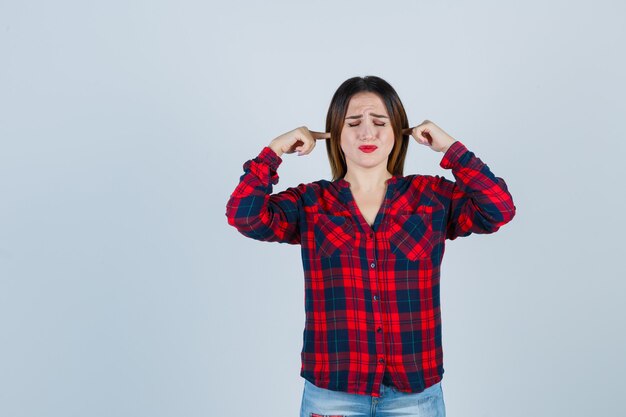 Retrato de joven hermosa mujer tapando los oídos con los dedos en camisa casual y mirando molesto vista frontal