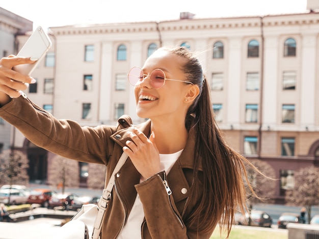 Retrato de joven hermosa niña sonriente en jeans y chaqueta hipster de verano. Modelo tomando selfie en smartphone. Mujer haciendo fotos en la calle. En gafas de sol