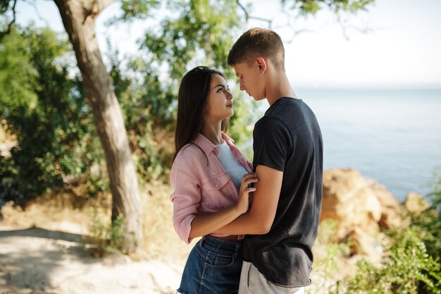 Retrato de una joven y hermosa pareja de pie y mirándose soñadoramente mientras pasan tiempo juntos con vistas al mar en el fondo