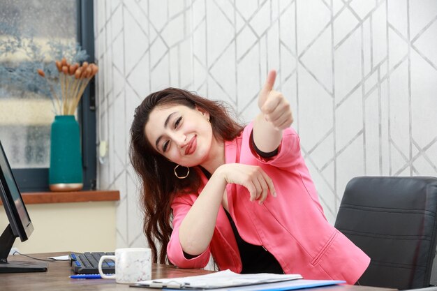 Retrato de una joven hermosa sentada en la oficina y gesticulando con el pulgar hacia arriba Foto de alta calidad