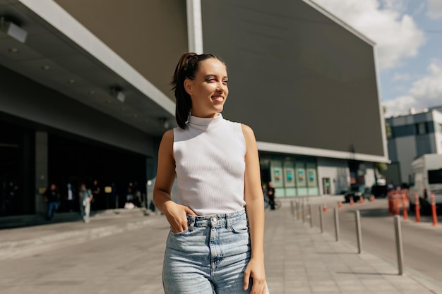 Retrato de una joven hermosa sonriendo mientras mira a un lado escondiéndose del sol al aire libre Una mujer de pelo largo con maquillaje brillante lleva pantalones cortos de mezclilla blancos