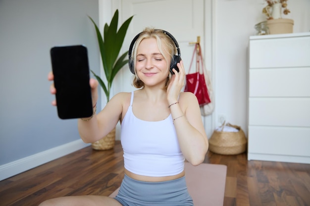 Foto gratuita retrato de una joven hermosa sonriente mostrando la pantalla de su teléfono inteligente con auriculares mirando