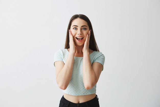 Retrato de joven hermosa sorprendida con la boca abierta sonriendo con las manos en las mejillas.