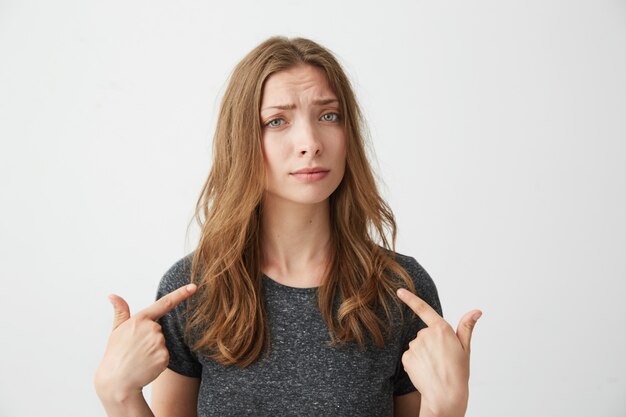 Retrato de joven hermosa sorprendida señalando con el dedo a sí misma.