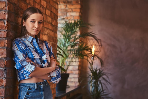 Retrato de una joven hipster vestida con una camisa y pantalones vaqueros cruzados de brazos mientras se apoyaba en una mesa en una habitación con interior de loft.