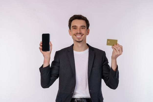 Retrato de un joven hombre de negocios feliz de pie mostrando un teléfono celular móvil y sosteniendo una tarjeta bancaria de crédito aislada en un estudio de fondo de color blanco