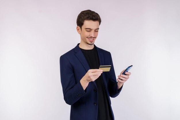 Retrato de un joven hombre de negocios feliz de pie usando un teléfono celular móvil y sosteniendo una tarjeta bancaria de crédito aislada en un estudio de fondo de color blanco