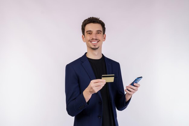 Retrato de un joven hombre de negocios feliz de pie usando un teléfono celular móvil y sosteniendo una tarjeta bancaria de crédito aislada en un estudio de fondo de color blanco