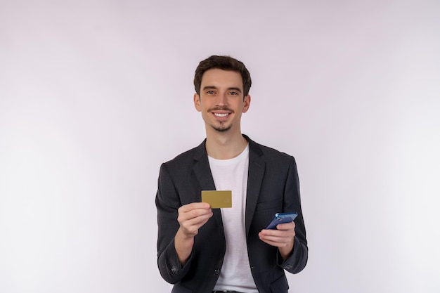 Retrato de un joven hombre de negocios feliz de pie usando un teléfono celular móvil y sosteniendo una tarjeta bancaria de crédito aislada en un estudio de fondo de color blanco