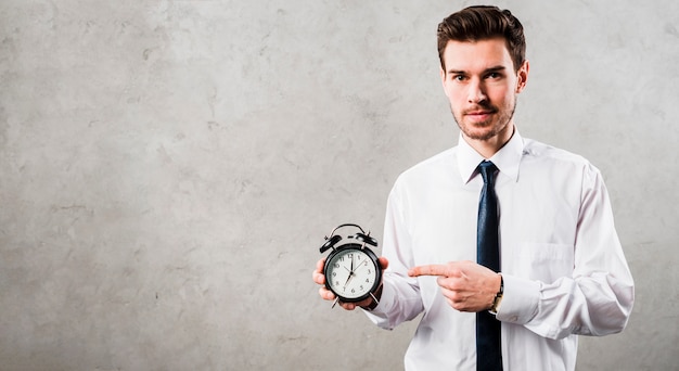 Retrato de un joven hombre de negocios que señala en el reloj de alarma negro que se opone al muro de cemento gris