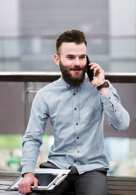 Retrato de un joven hombre de negocios sentado en el banco con portapapeles y tableta digital hablando por teléfono móvil