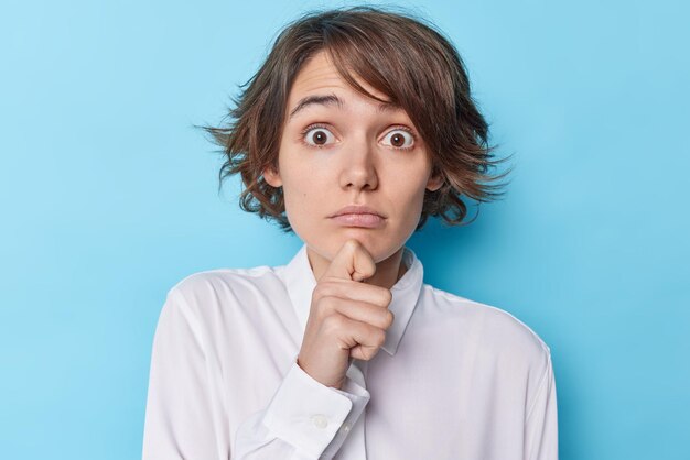 El retrato de una joven impresionada sostiene la barbilla mira fijamente con los ojos reventados y escucha noticias impactantes, tiene un peinado moderno, usa una camisa blanca aislada sobre un fondo azul y se siente abrumado. reacción humana