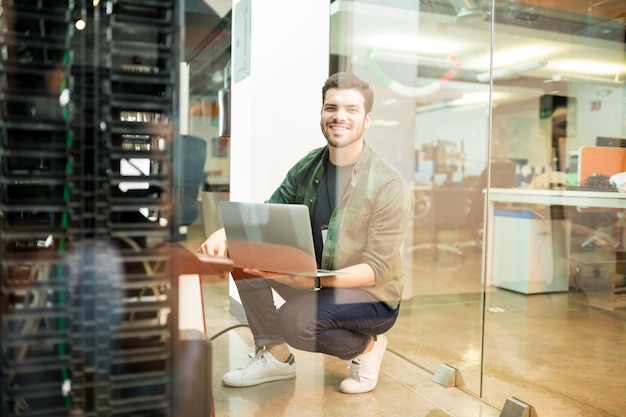 Foto gratuita retrato de un joven ingeniero de redes masculino feliz con una laptop en la mano trabajando en un centro de datos.