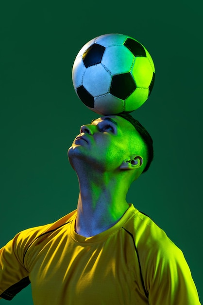 Foto gratuita retrato de un joven jugador de fútbol con una pelota en la cabeza posando aislado sobre un fondo verde de estudio con luz de neón