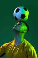 Foto gratuita retrato de un joven jugador de fútbol con una pelota en la cabeza posando aislado sobre un fondo verde de estudio con luz de neón