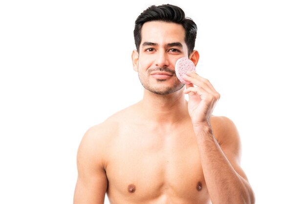 Retrato de un joven latino limpiando su cara con una esponja facial sobre fondo blanco.